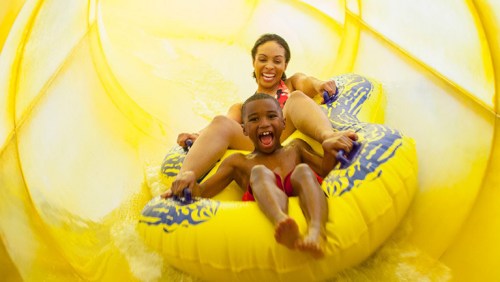 A woman and a child joyfully slide down a yellow water slide together, both smiling and having fun.