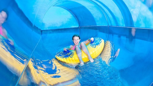 A child smiles while riding a yellow inner tube down a blue water slide.