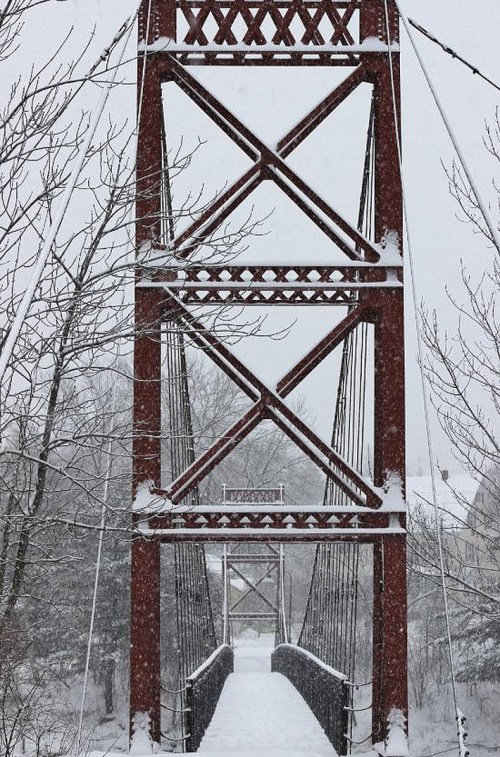 A snow-covered bridge with a red metal frame, surrounded by trees in a winter landscape.