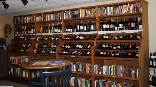 A wooden shelf filled with wine bottles and books, with a small table and chair in front.