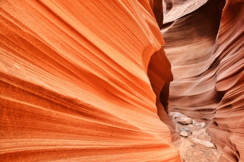 A narrow canyon with vibrant orange and red rock formations, showcasing smooth, wavy textures and a rocky floor.