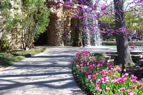 A scenic garden path lined with pink tulips, leading to a stone structure and a waterfall surrounded by blooming trees.