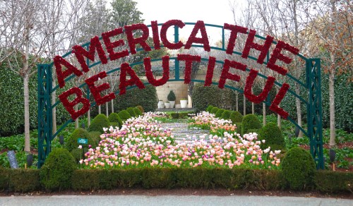 A floral archway reading "America the Beautiful" above a colorful garden of tulips and greenery.