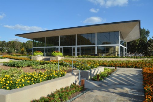Modern glass building surrounded by vibrant flower beds and greenery under a clear blue sky.