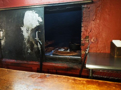 A smoky barbecue pit with meat on a grill, set against a rustic red wall.