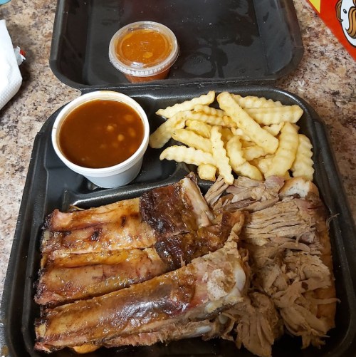 A takeout container with ribs, pulled pork, crinkle fries, and a side of barbecue sauce and baked beans.