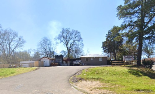 A gravel road leads to a building with a sign, surrounded by trees and a clear blue sky.