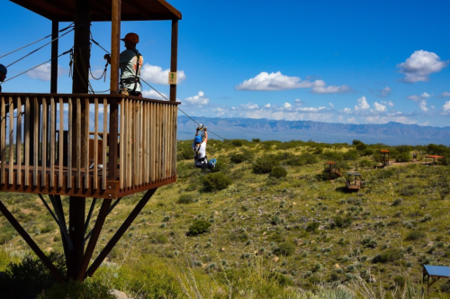 A person zip-lining from a wooden platform over a green landscape with mountains and blue sky in the background.