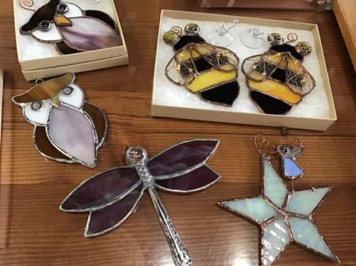 Colorful stained glass ornaments including an owl, dragonfly, star, and bee earrings displayed on a wooden surface.