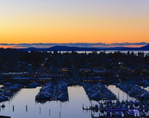 Sunset over a marina, with boats docked and reflections on the water, surrounded by distant hills and a colorful sky.