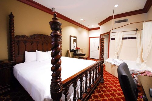 A luxurious hotel room featuring a wooden four-poster bed, a sitting area, and a bathroom with a tub.