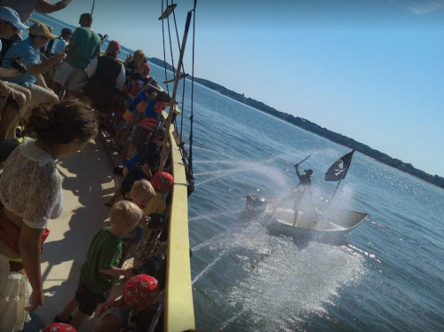 A pirate on a small boat sprays water while a crowd watches from a larger vessel on a sunny day.