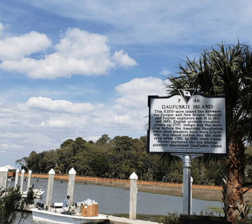 A historical marker about Daufuskie Island near a dock with boats, surrounded by trees and a blue sky.