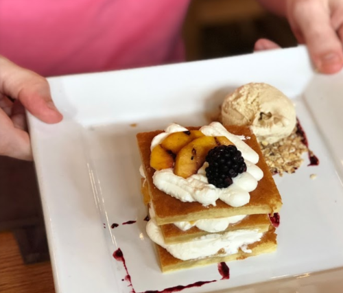 A person holds a plate with layered dessert, featuring cream, fruit, and a scoop of ice cream on the side.