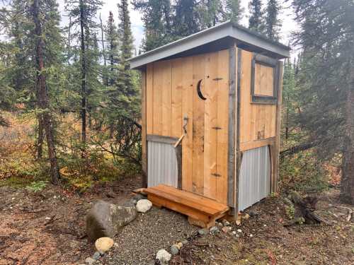 A rustic wooden outhouse surrounded by trees in a natural setting, featuring a simple door and a wooden step.