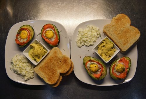 Two plates with baked avocado halves topped with eggs, toast, and bowls of hummus and cottage cheese.