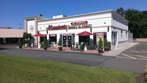 A bakery storefront with outdoor seating, red umbrellas, and a sign reading "De Pasquale Bakery & Pastry."