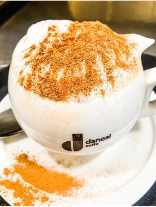 A close-up of a frothy coffee topped with a dusting of cinnamon, served in a white cup on a saucer.