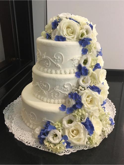 Three-tiered white wedding cake adorned with white and blue flowers, featuring elegant decorative swirls.