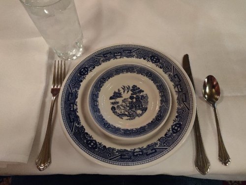 A table setting with a blue and white patterned plate, silver utensils, and a glass of water on a white tablecloth.