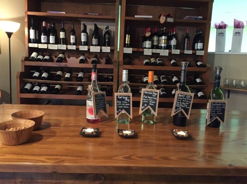 A wooden bar with various bottles of wine and spirits, alongside small bowls of snacks and a wine rack in the background.