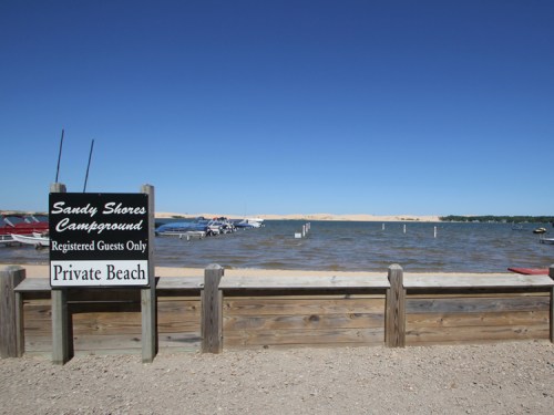 Sign for Sandy Shores Campground by a lake with boats and a sandy dune in the background under a clear blue sky.