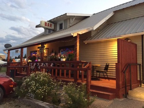 A cozy bar with a wooden porch, outdoor seating, and TVs, set against a twilight sky.