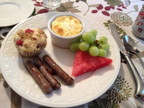 A plate with sausages, a muffin, a baked egg dish, grapes, and watermelon, set on a floral tablecloth.