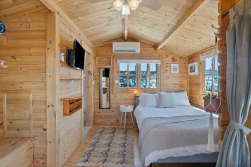 Cozy wooden interior of a small cabin with a bed, TV, and large windows overlooking a marina.