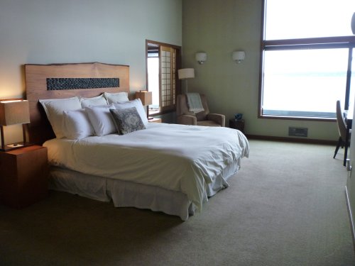 A spacious bedroom featuring a large bed, bedside tables, a chair, and a window with a view. Soft, neutral colors.