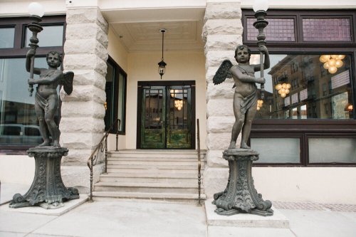 Two angelic statues holding lamps stand on either side of a grand entrance with steps and large windows.