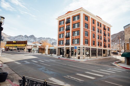 A brick building with balconies stands at a street corner, surrounded by mountains and shops under a clear sky.