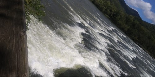 A river with white water rapids flows through a lush green landscape under a partly cloudy sky.