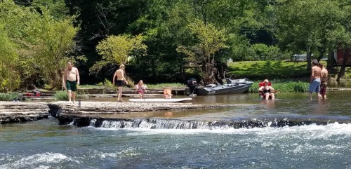 People enjoying a sunny day by the river, with some swimming, paddleboarding, and relaxing on the shore.