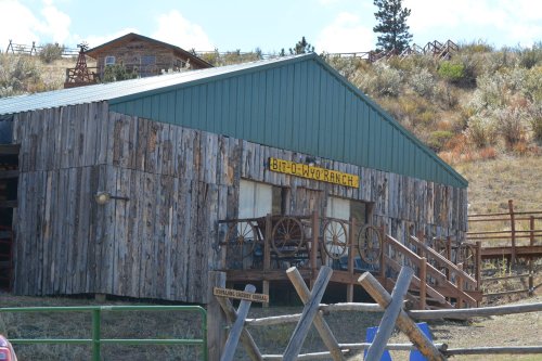 A rustic wooden building with a green roof, featuring a sign that reads "Bit-O-W Ranch" and decorative wagon wheels.