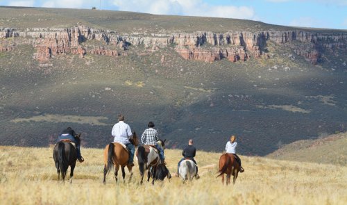 A group of horseback riders traverses a grassy landscape with rocky cliffs in the background under a blue sky.