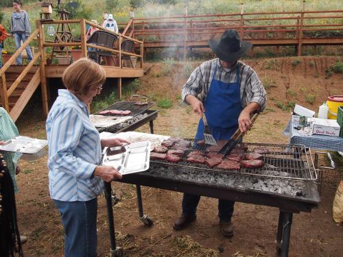 A person in a cowboy hat grills burgers outdoors while another holds a tray, surrounded by a rustic setting.