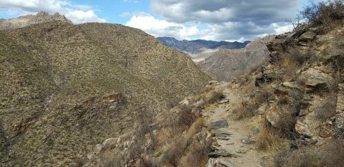 A rocky trail winds through arid mountains under a partly cloudy sky.