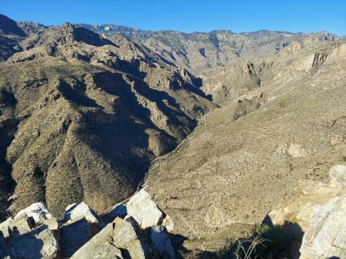 A panoramic view of rugged mountains and valleys under a clear blue sky, showcasing rocky terrain and shadows.