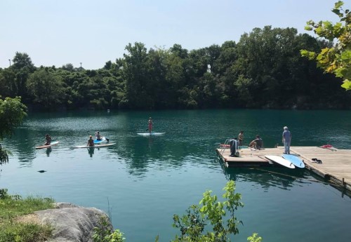 People paddleboarding on a calm lake with a wooden dock and lush greenery in the background.