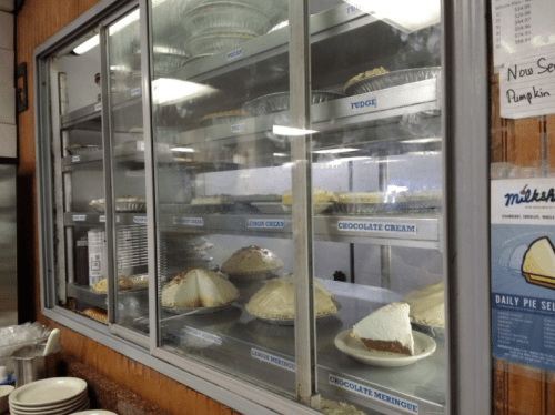 A display case filled with various pies, including chocolate cream, lemon cream, and fudge, in a cozy diner setting.