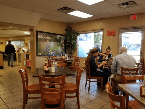A cozy restaurant interior with diners at tables, a mural of flowers, and a person walking towards the counter.