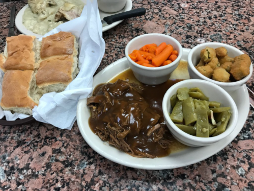 A plate of pulled pork with gravy, green beans, carrots, fried okra, and biscuits on a pink speckled table.