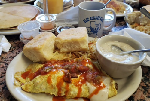 A plate with an omelet topped with salsa, two biscuits, hash browns, and a bowl of gravy on a restaurant table.