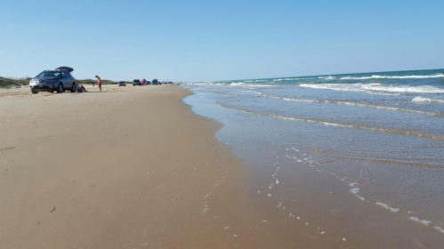 A sandy beach with gentle waves, cars parked along the shore, and a clear blue sky in the background.