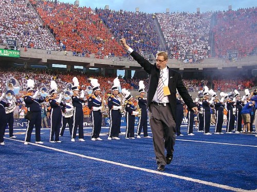 A conductor leads a marching band on a blue football field, surrounded by fans in orange and blue stands.