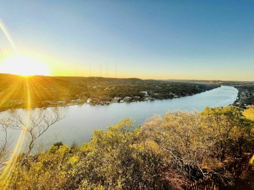 Sunset over a serene river, surrounded by lush greenery and distant hills, with radio towers visible on the horizon.