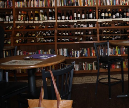A cozy wine bar with wooden shelves filled with bottles and books, featuring round tables and black chairs.