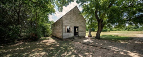 A rustic wooden house surrounded by trees and grass, with a clear blue sky in the background.