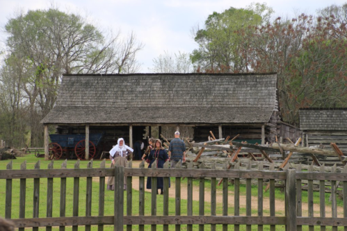 A historic farm with a wooden building and visitors walking along a path, surrounded by a wooden fence and greenery.